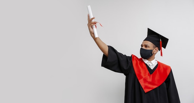 Glücklicher Absolventenjunge, er ist in einem schwarzen Mörtelbrett mit roter Quaste und einer Gesichtsmaske im Gewand und schaut auf das Diplom in seiner Hand Heller Hintergrundkopierraum