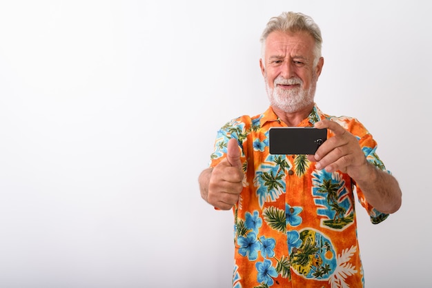 glücklicher älterer bärtiger Touristenmann, der lächelt, während er Daumen aufgibt und Foto mit Handy auf Weiß macht
