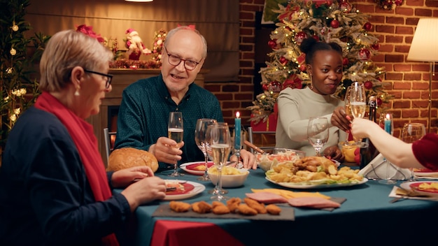 Glücklicher älterer Mann, der mit seiner Frau spricht, während er den Weihnachtstisch mit nahen Familienmitgliedern genießt. Multiethnische festliche Menschen, die traditionelle Winterferien feiern, während sie hausgemachtes Essen essen.