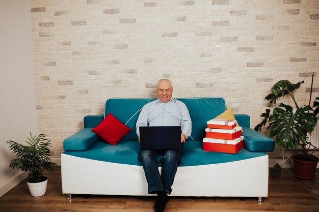 Glücklicher älterer Mann, der zu Hause auf blauem Sofa sitzt und Laptop verwendet. Remote-Arbeit online. Senior Geschäftsmann im Home Office.