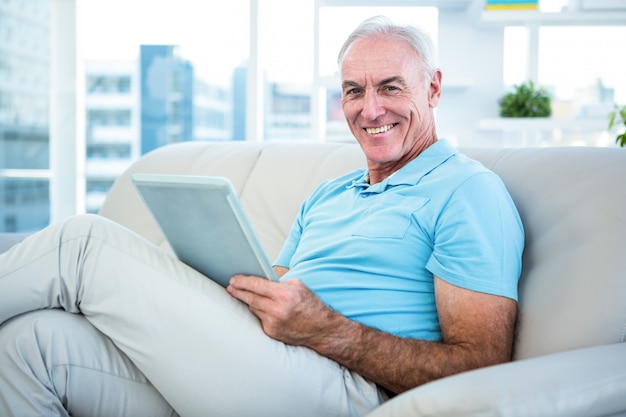 Glücklicher älterer Mann, der zu Hause auf Sofa mit digitaler Tablette sitzt