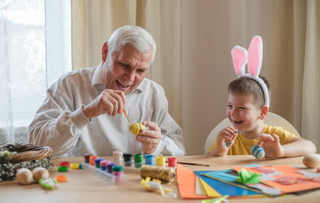 Glücklicher älterer Manngranvater, der Ostern mit Enkel vorbereitet. Porträt des lächelnden Jungen mit Hasenohren malte bunte Eier für Ostern