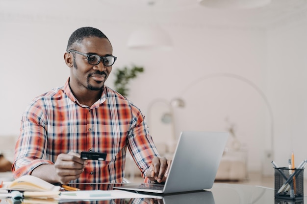 Glücklicher afrikanisch-amerikanischer Mann mit Brille kauft online mit Laptop und Kreditkarte