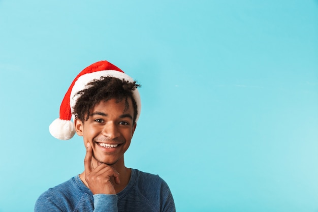 Glücklicher afrikanischer Mann, der roten Weihnachtshut trägt, der über blauer Wand lokalisiert wird