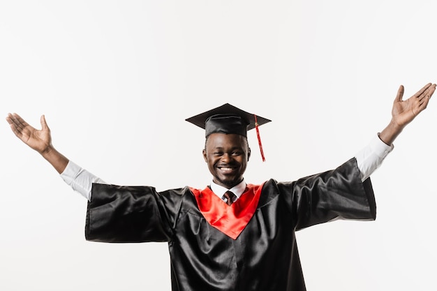 Glücklicher afrikanischer Student in schwarzem Abschlusskleid und Mütze hebt seine Hände über dem Kopf auf weißem Hintergrund Der Absolvent des afrikanischen Mannes absolviert das College und feiert akademische Leistungen