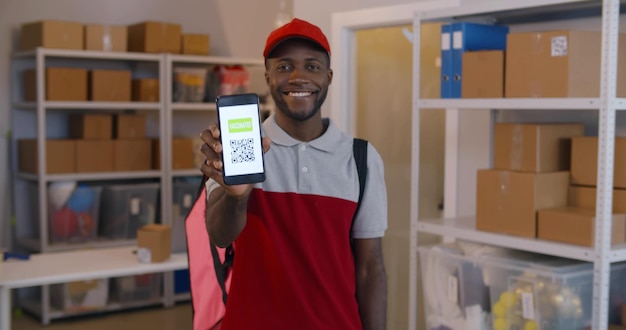 Glücklicher afroamerikanischer Lieferbote lächelt in die Kamera und zeigt Mobiltelefon mit geimpftem QR-Code