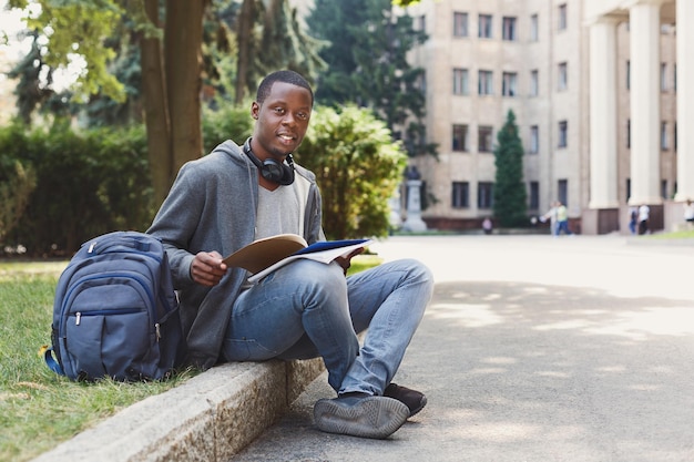 Glücklicher afroamerikanischer Student, der mit Notebook auf dem Universitätscampus sitzt. Mann, der draußen mit Schreibheft studiert. Bildung, Inspiration und Fernarbeitskonzept.