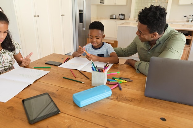 Glücklicher afroamerikanischer Vater mit Tochter und Sohn, die zu Hause lächelnd Hausaufgaben machen. Familienleben, gemeinsames Arbeiten zu Hause.