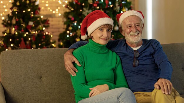 Glücklicher alter Mann in Weihnachtsmütze, der die Kamera mit einem Lächeln anschaut und eine reife Frau umarmt, während er abends auf der Couch auf unscharfem Hintergrund der Weihnachtsdekoration sitzt