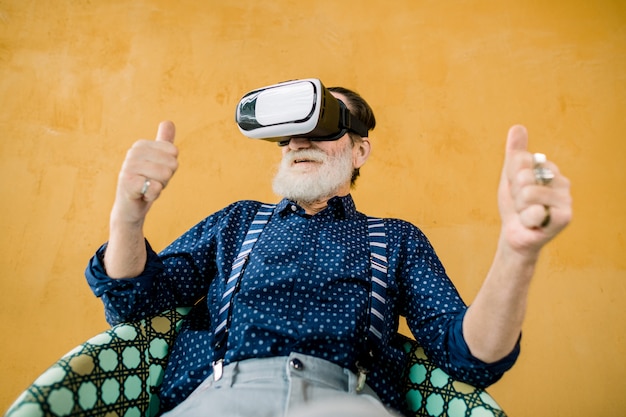 glücklicher alter Mann mit gepflegtem Bart, der seine Daumen zeigt, während er Virtual-Reality-Brillen zum Ansehen von Filmen verwendet. Studioaufnahme auf gelbem Hintergrund. VR-Technologien
