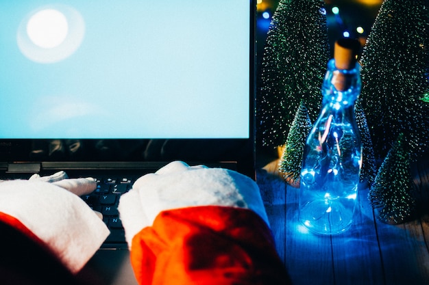 Glücklicher alter Weihnachtsmann mit Hut, der Geschenkbox mit Laptop-Computer hält, der spät am frohen Weihnachtsabend am Tisch der Werkstatt sitzt. E-Commerce-Website Weihnachtszeit Urlaub Online-Shopping E-Commerce-Verkauf