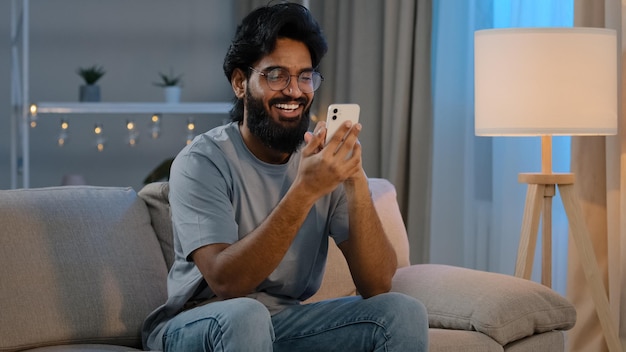 Glücklicher arabischer indischer männlicher tausendjähriger mann, der zu hause moderne smartphone-sms-nachricht hält