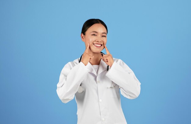 Glücklicher asiatischer Arzt in Uniform, der mit beiden Händen auf ihr Lächeln zeigt