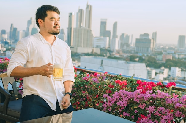 Glücklicher asiatischer Geschäftsmann, der lächelt, während er Bier am Dachrestaurant trinkt