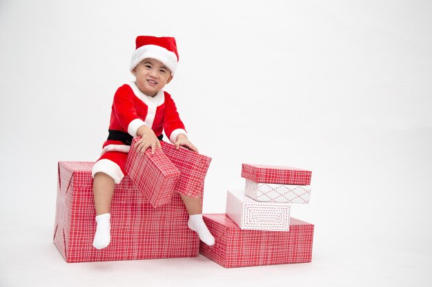 Glücklicher asiatischer junge mit weihnachtsmannanzug, der geschenkbox hält und auf roter geschenkbox lokalisiert auf weißer wand sitzt