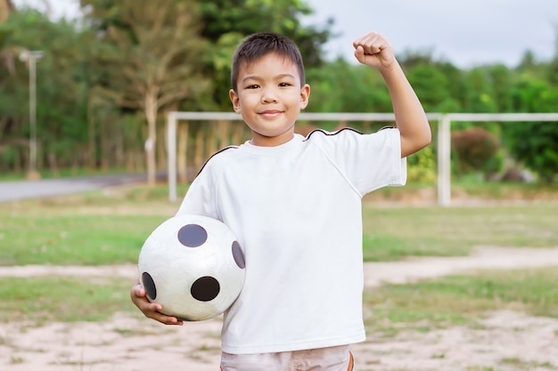 Glücklicher asiatischer Kinderjunge, der ein Fußballspielzeug in seinen Händen spielt und hält Er trägt weißes Hemd auf dem Feldspielplatz Glücklicher und lächelnder Junge