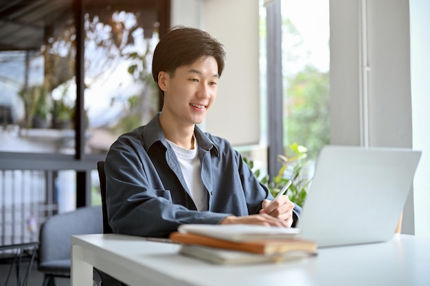 Glücklicher asiatischer männlicher Student, der Hausaufgaben im Café mit Laptop und Tablet macht