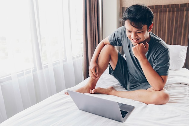 Glücklicher asiatischer Mann arbeitet mit seinem Laptop auf seinem Bett Konzept eines erfolgreichen Lebensstils für Freiberufler