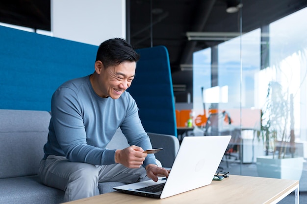 Glücklicher asiatischer Mann, der online in einem modernen Büro auf einem Laptop einkauft, lächelt und sich über das ausgewählte Produkt freut, das eine Kreditkarte hält