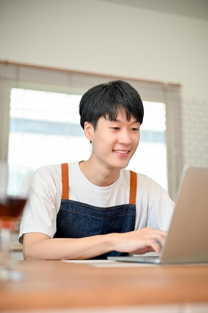 Glücklicher asiatischer Mann in Schürze mit Laptop an seiner Küchentheke auf der Suche nach Lebensmittelrezepten