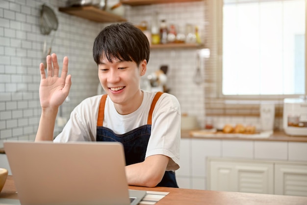 Glücklicher asiatischer Mann in Schürze mit Laptop, der einen Online-Videoanruf über Laptop in der Küche hat