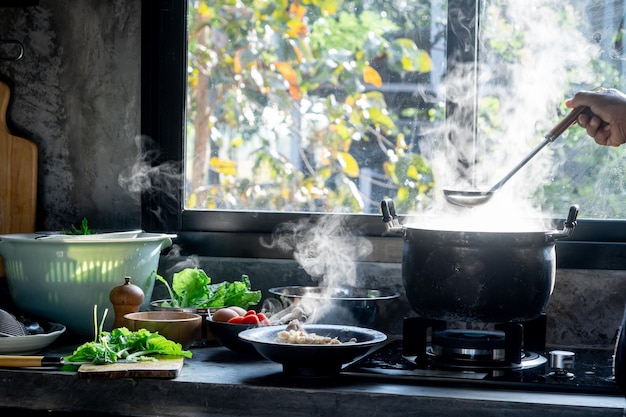 Foto glücklicher asiatischer mann kocht, während er den schöpflöffel in einem großen topf benutzt. das wasser kocht und die dampfmasse spiegelt sich im morgenlicht in der küche. küchenutensilien und werkzeug zu hause