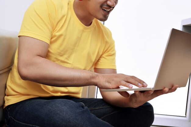 Glücklicher asiatischer Mann mit dem Laptop, der auf der Couch sitzt