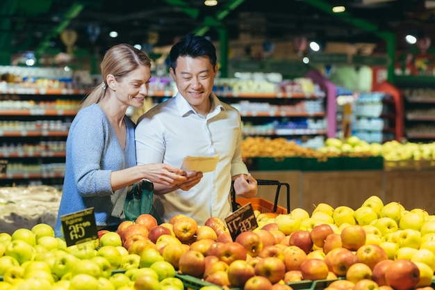 Glücklicher asiatischer Paarfamilienmann und -frau der gemischten Rasse, die Obst oder Gemüse im Lebensmittelgeschäft wählen