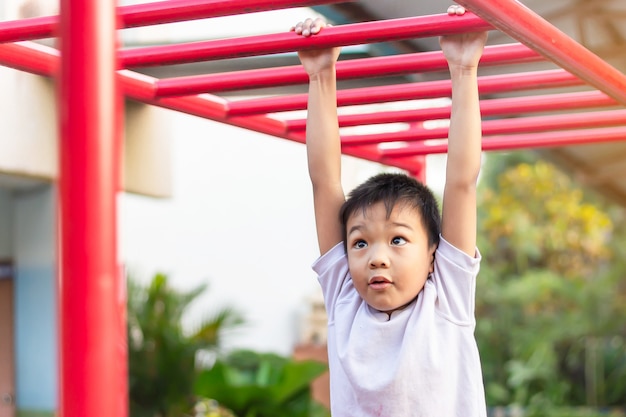 Glücklicher asiatischer studentenkindjunge, der von einer stahlstange am spielplatz spielt und hängt.