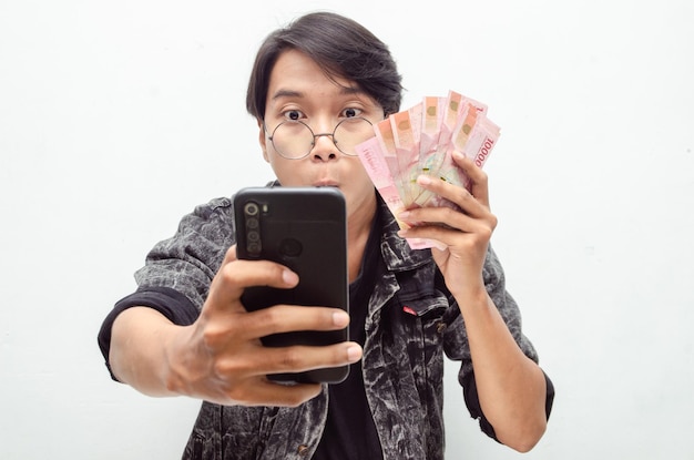 Glücklicher attraktiver indonesischer junger mann schockiert glücklich, während er rupiah-papiergeld und telefon hält.