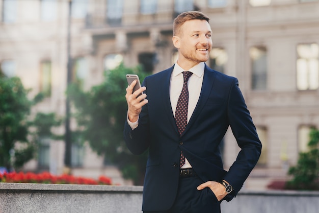Glücklicher attraktiver männlicher Finanzier in der eleganten Abnutzung, unter Verwendung des modernen Smartphone
