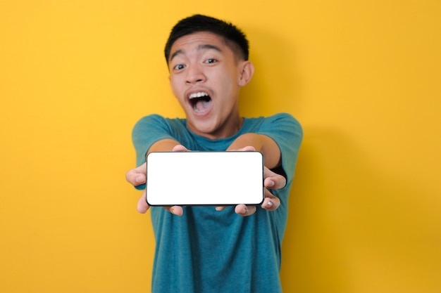 Glücklicher aufgeregter junger asiatischer Mannschock, der weißen Telefonbildschirm an der Kamera zeigt, lokalisiert auf gelbem Hintergrund