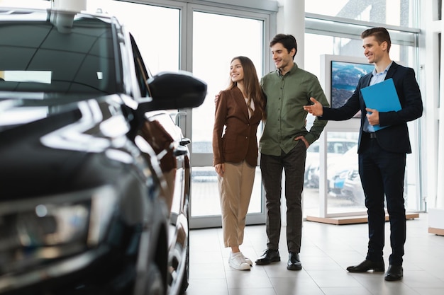 Glücklicher Autoverkäufer, der jungen Paaren im Autohaus neue Autos zeigt