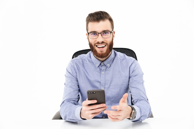 Glücklicher bärtiger, eleganter Mann mit Brille, der Smartphone benutzt und beim Sitzen am Tisch schaut