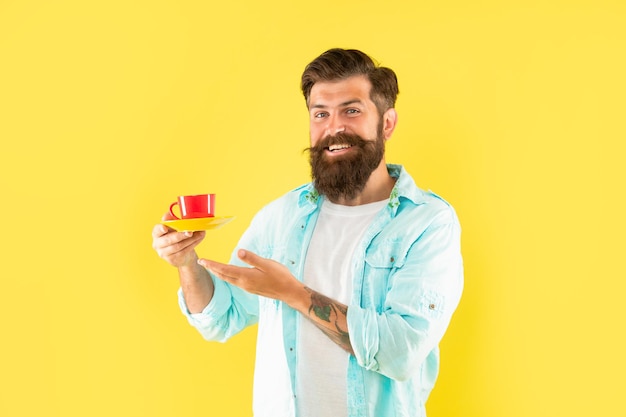 Glücklicher bärtiger Kerl, der Morgenkaffee auf gelbem Hintergrund, Getränk präsentiert