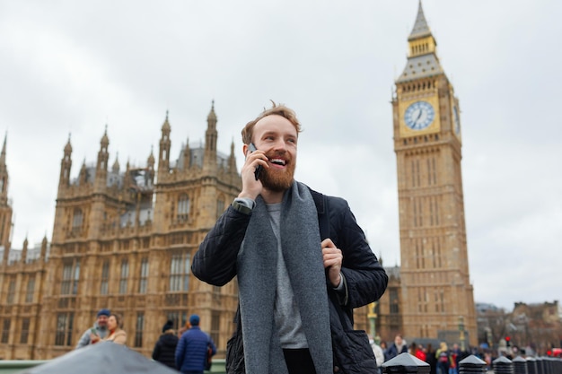 Glücklicher bärtiger Mann, der am Telefon spricht, während er auf dem Hintergrund des Big Ben-Turms steht