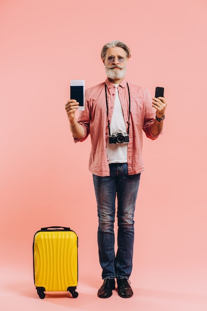 Glücklicher bärtiger Mann in der Sonnenbrille, die ein Handy und einen Pass mit Tickets auf Rosa hält