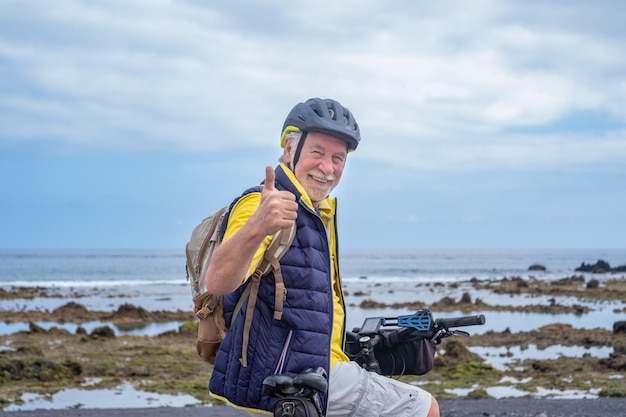 Glücklicher bärtiger Radfahrer, älterer Mann am Meeresstrand, der einen Rucksack mit Helm trägt, der mit einem Elektrofahrrad läuft, das ein Ok-Zeichen gestikuliert
