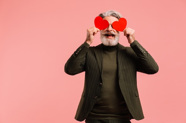 Glücklicher bärtiger und reifer Mann des Schnurrbartes im khakifarbenen Anzug, der zwei rote Herzen nahe Augen hält, die Valentinstag isolierten rosa Hintergrund begrüßen.