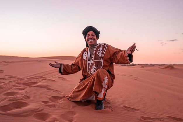 Glücklicher Beduinenmann in traditioneller Kleidung, der Zigarette hält, während er auf Sand posiert