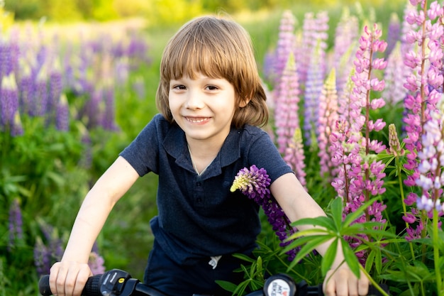 Glücklicher blonder Junge in einem Feld mit Blumenlupinen auf einem Fahrrad.