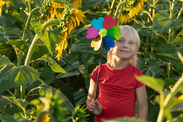 Glücklicher blonder Vorschulkind mit einer Spielzeugwindmühle in seinen Händen steht auf einer grünen Wiese