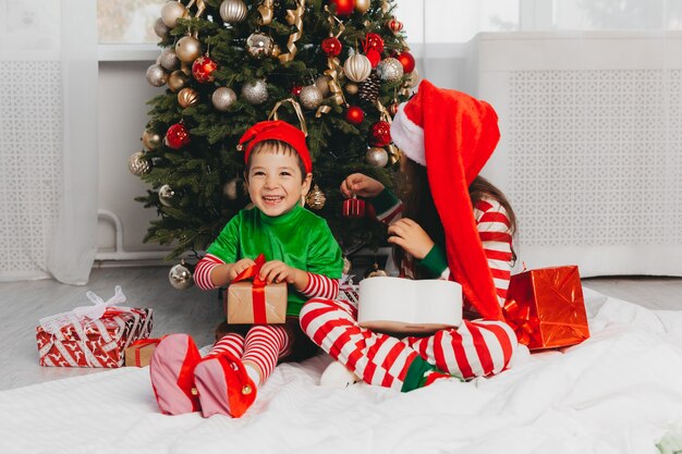 Glücklicher Bruder und Schwester, die als Weihnachtsmann verkleidet sind, sitzen zu Hause im Wohnzimmer mit Geschenken in der Nähe des Weihnachtsbaums. Weihnachten