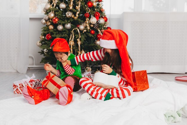 Glücklicher Bruder und Schwester, die als Weihnachtsmann verkleidet sind, sitzen zu Hause im Wohnzimmer mit Geschenken in der Nähe des Weihnachtsbaums. Weihnachten