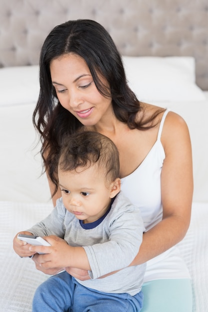 Glücklicher Brunette, der ihr Baby hält und Smartphone im Schlafzimmer verwendet