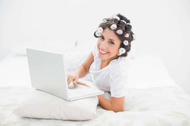 Glücklicher Brunette in den Haarrollen, die auf ihrem Bett unter Verwendung ihres Laptops liegen