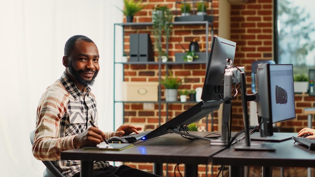 Glücklicher Ersteller von Inhalten, der eine Softwareschnittstelle auf einem Touchscreen-Display mit Tablet und Stift am Schreibtisch entwickelt. Afroamerikaner, der an der Produktion von Infografiken im Agenturstudio arbeitet.