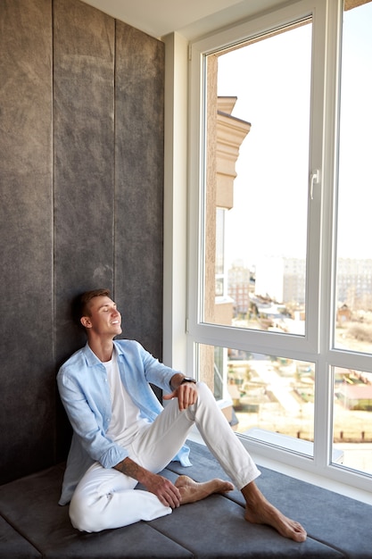 Glücklicher erwachsener kaukasischer Mann sitzt nahe Panoramafenster und kühlt