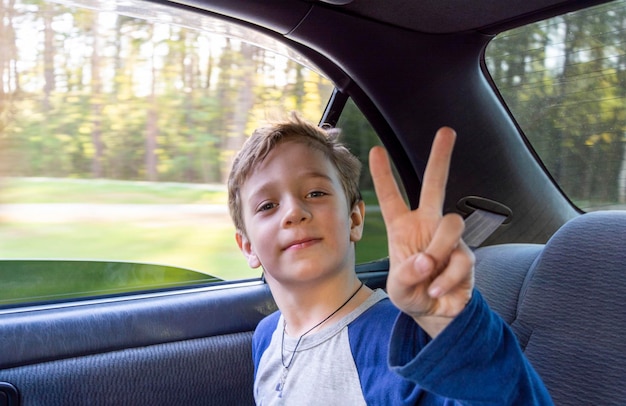 Glücklicher europäischer Junge fährt im Auto auf dem Rücksitz, das Kind zeigt das Victory-Zeichen