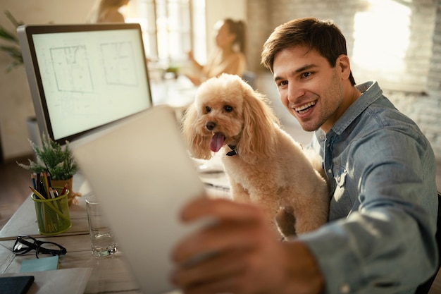 Glücklicher Freiberufler, der Spaß hat, während er Touchpad benutzt und Selfie mit seinem Hund im Büro macht